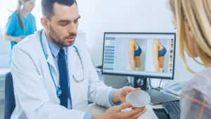 A surgeon holding a breast implant while a patient views breast images on a screen